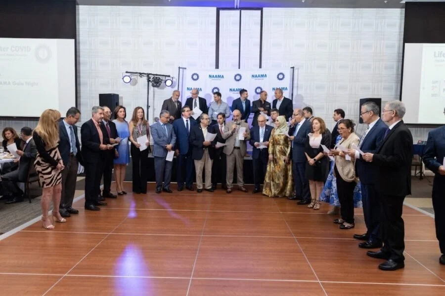 Group of diverse professionals standing in a circle at a conference or awards ceremony, with a stage and banners in the background, and some holding certificates or papers.