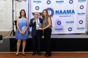 Three women standing on a stage, one in the middle holding an award and a man beside her holding the award with a NAAMA backdrop behind them.