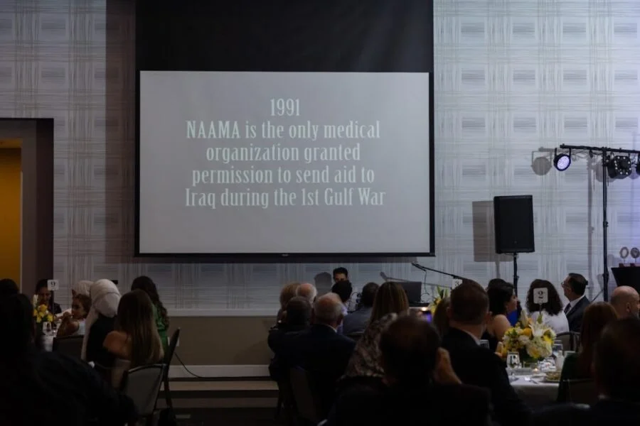 A conference room with a large screen displaying text about NAAMA and the Gulf War, surrounded by seated attendees.