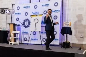 A man in a suit speaking on stage at a NAAMA event, with a backdrop displaying the NAAMA logo and a question mark.