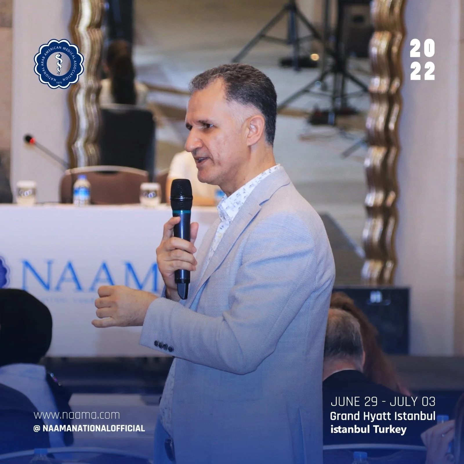 A man with short gray hair and light skin wearing a light-colored suit jacket and a patterned shirt speaks into a black microphone at a conference at the Grand Hyatt Istanbul in Istanbul, Turkey, held from June 29 to July 3 in 2022. The background sh