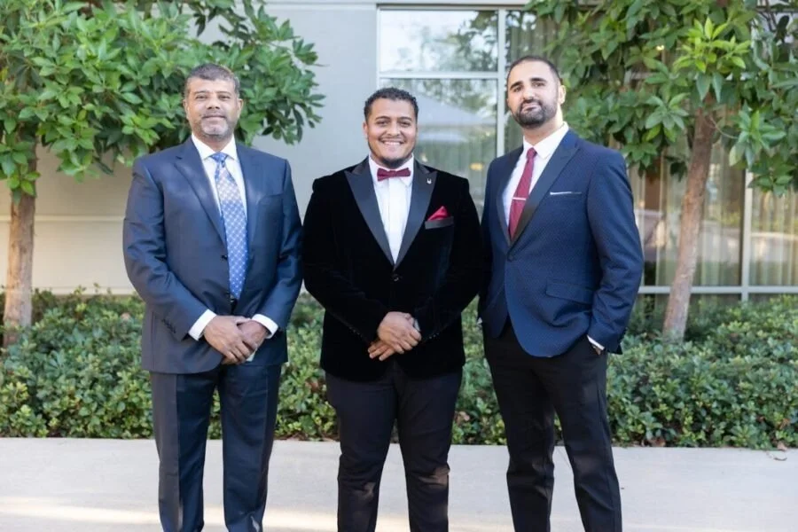 Three men in formal suits standing outdoors in front of bushes and a building, smiling at the camera.