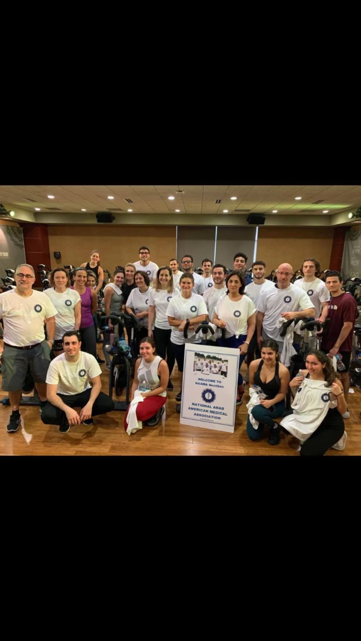 Group photo of people in a fitness class at a gym, with a sign that reads 'Welcome to NAAMA NextGen, National Arab American Medical Association'