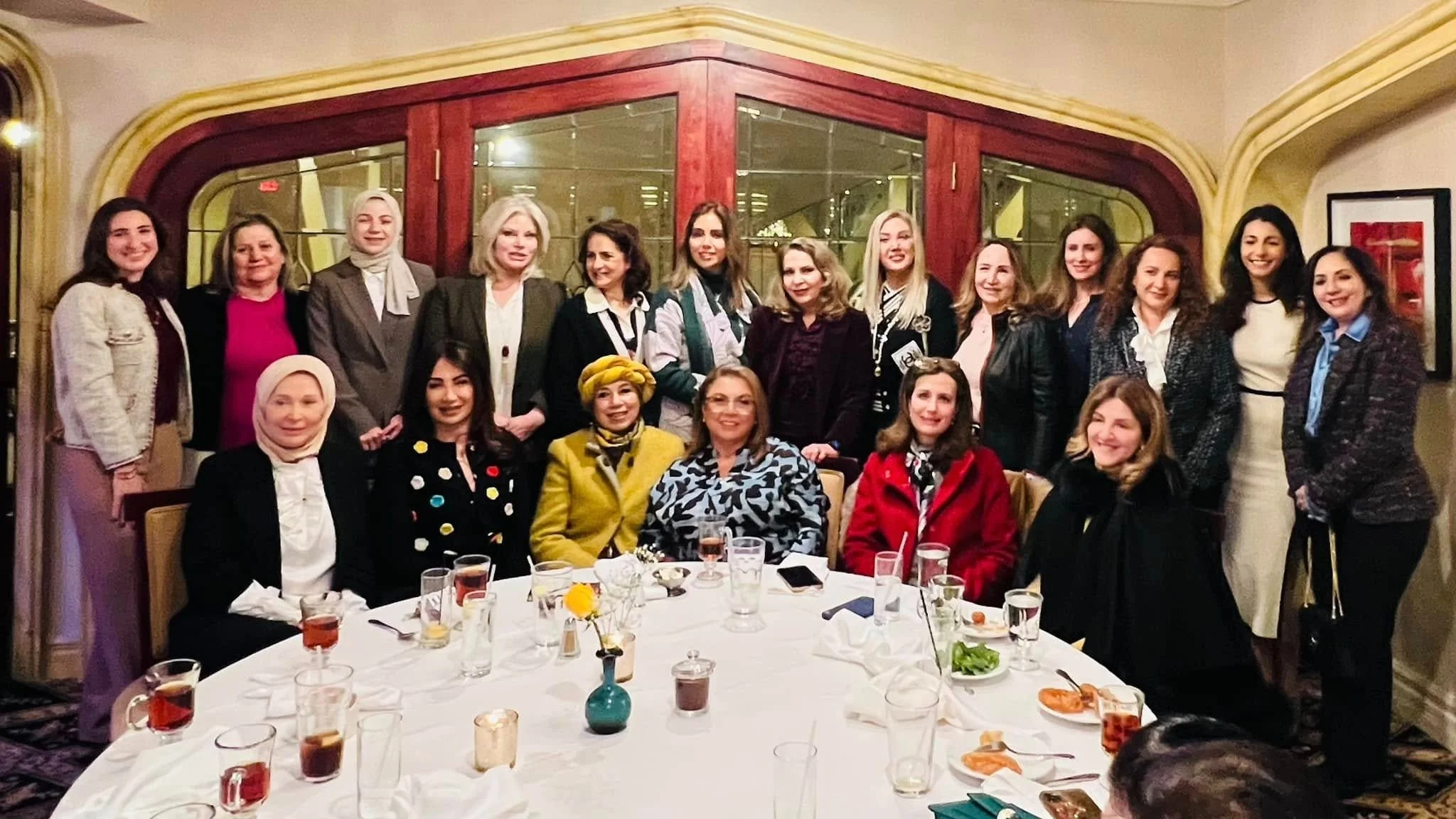 Group of women gathered around a table at a social gathering or dinner event, with some standing behind and others sitting in front. The table has glasses, plates, and small vases with flowers.