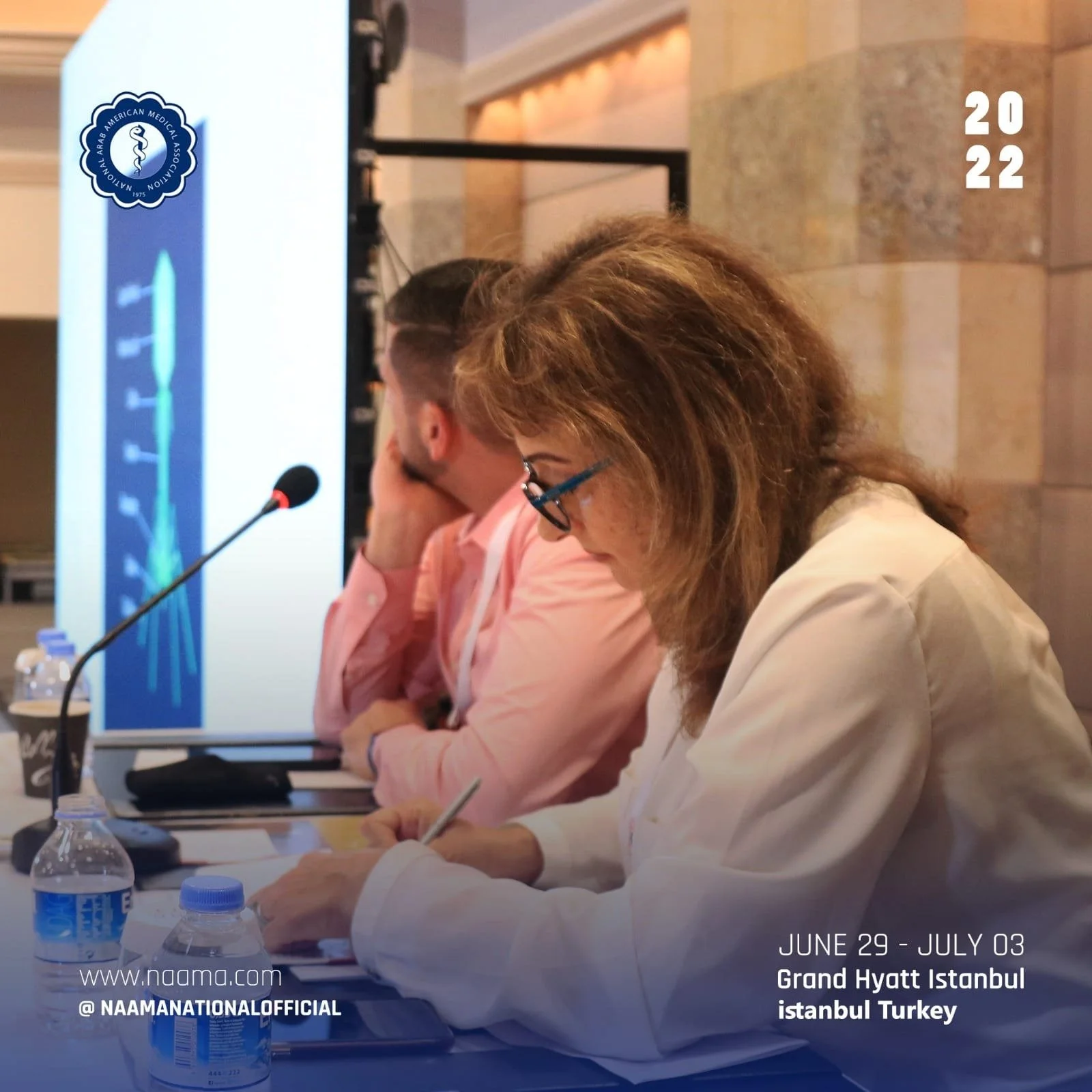 A group of people sitting at a long table during a conference or meeting, with microphones, bottled water, and notebooks in front of them. There is a presentation screen on the side displaying the NAAMA (National Arab American Medical Association) lo