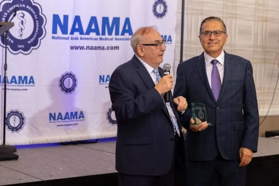 Two men in suits standing on a stage at a NAAMA event. One man is speaking into a microphone, and the other is holding a glass award. Behind them is a white banner with the NAAMA logo and text.