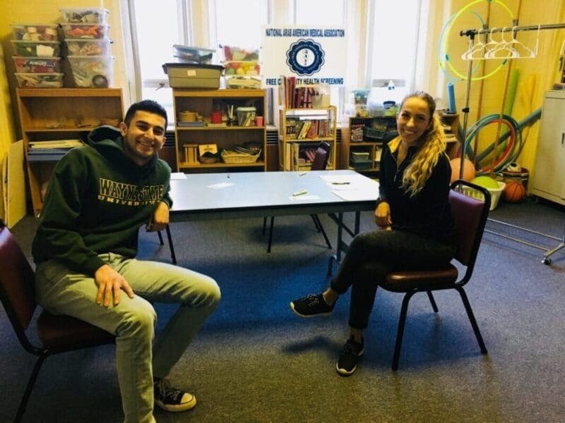 Two people sitting at a table in a room with sports equipment and bookshelves, smiling at the camera.