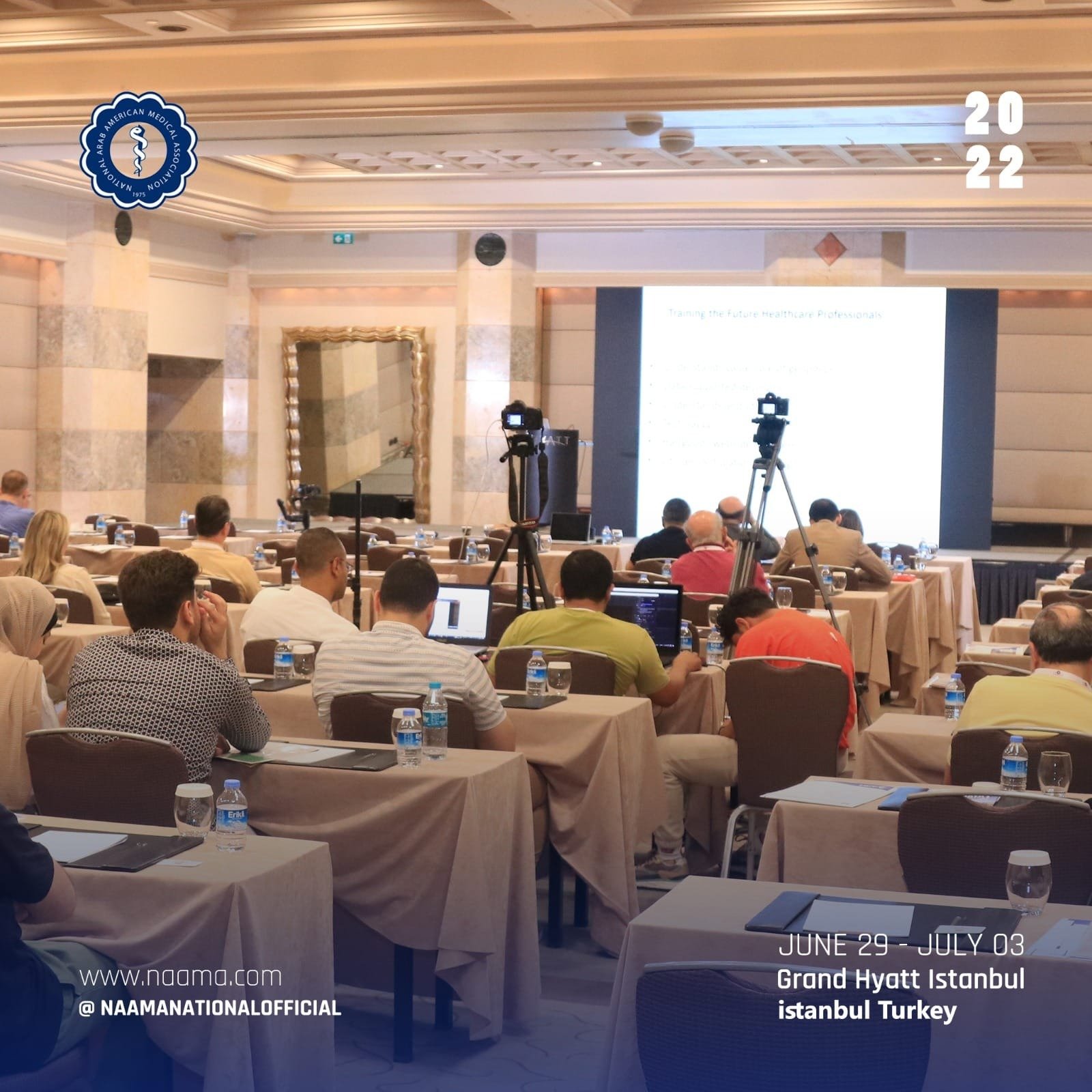 A conference room filled with attendees seated at tables, some using laptops, facing a large screen displaying a presentation. There are multiple cameras on tripods set up in the room. The room has elegant architecture with decorative moldings. The e