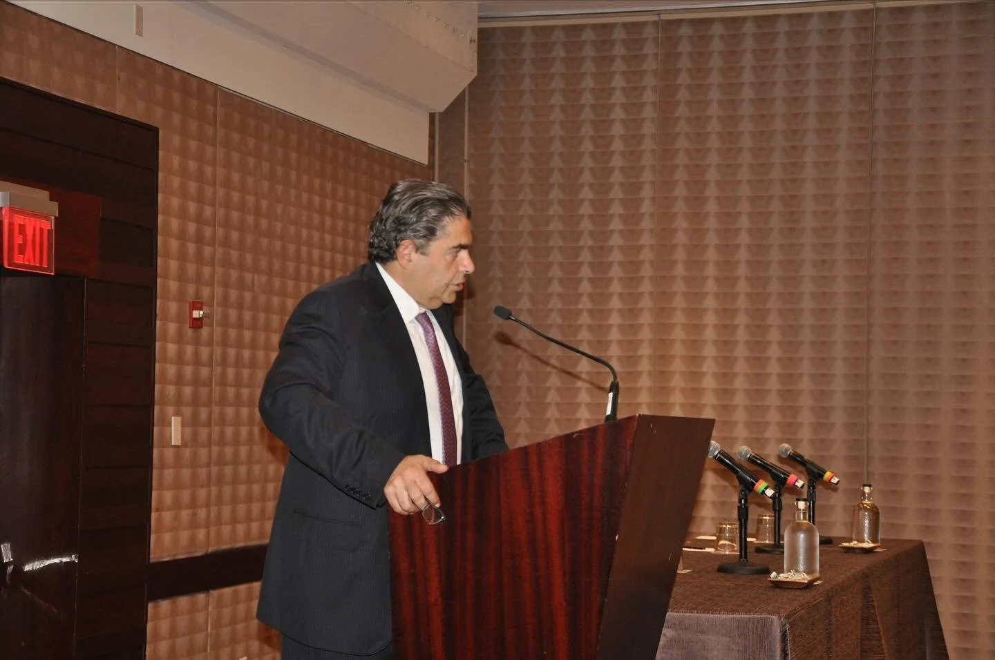 A man in a dark suit standing at a podium, speaking into a microphone, in a conference room with brown textured walls and a table with three microphones, glasses, and bottles on it.