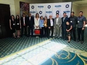 Group of people standing together in a conference room with a NAMM banner in the background.