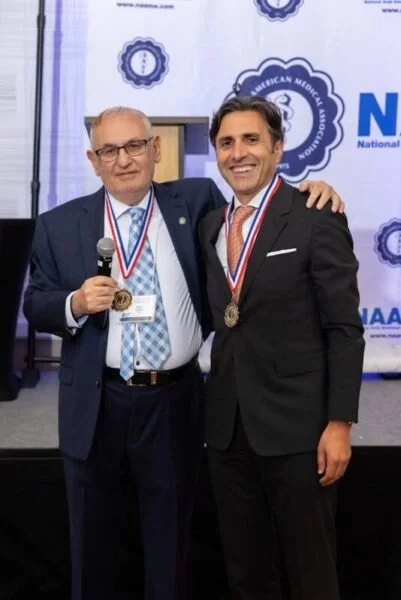 Two men in formal suits wearing medals around their necks, standing together at an event, smiling for the photo.