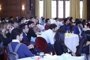 Audience of young adults attending a conference or seminar in a large room with wooden paneling and large windows.
