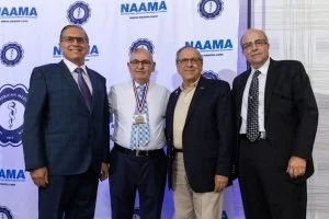 Four men in formal attire standing together at an NAAMA event with NAAMA banners in the background.