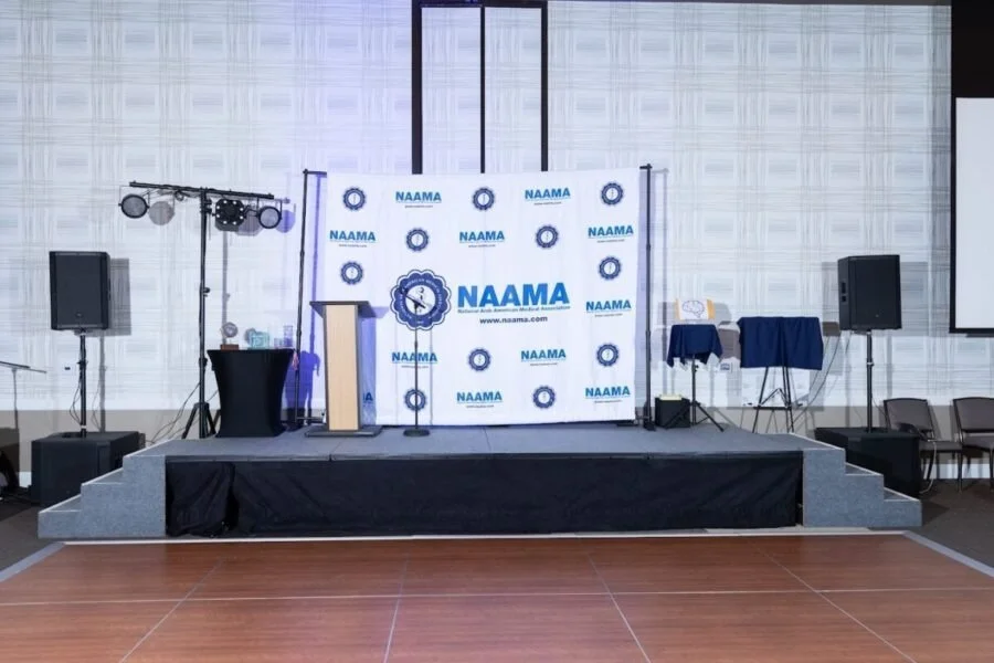 Event stage with NAAMA branding backdrop, podium, microphones, speakers, and lighting equipment in conference room.