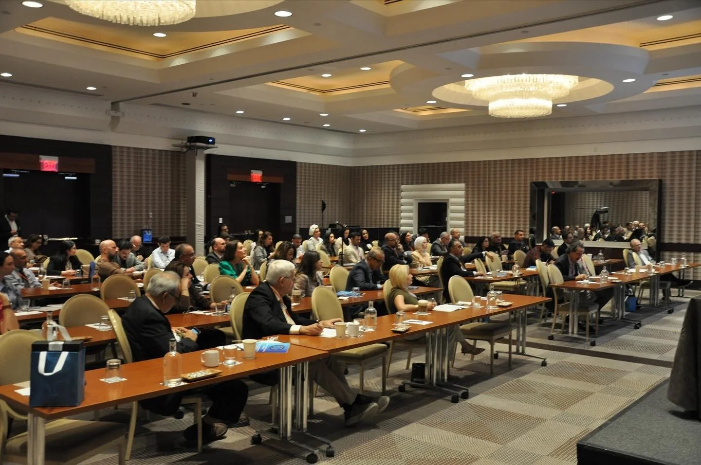 A large conference room filled with people sitting at long tables, listening to a presentation. The room has a modern design with a high ceiling and a chandelier.