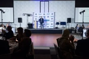 A person on stage speaking or performing at a conference, with large screens and audio equipment behind them. The audience sits at tables facing the stage in a dimly lit room.