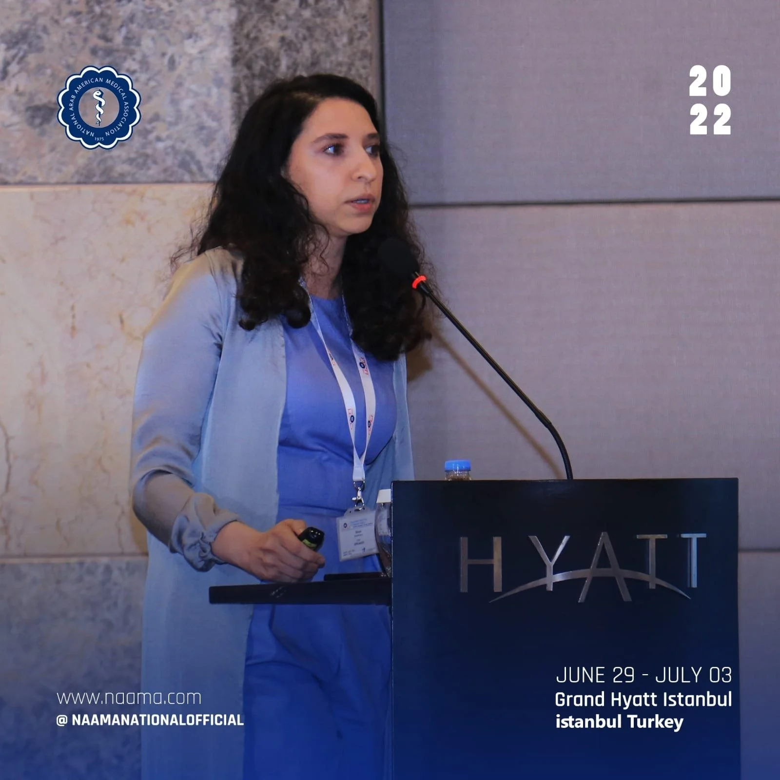 A woman with dark curly hair speaking at a conference at the Grand Hyatt Istanbul. She is wearing a blue dress and a light gray blazer, standing behind a podium with the Hyatt logo, with a microphone in front of her. The background includes a marble 