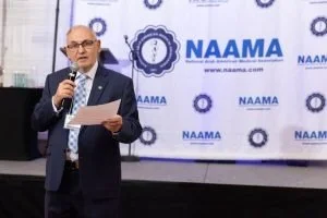 A man in a suit speaking into a microphone at a NAAMA event, with a NAAMA banner in the background.
