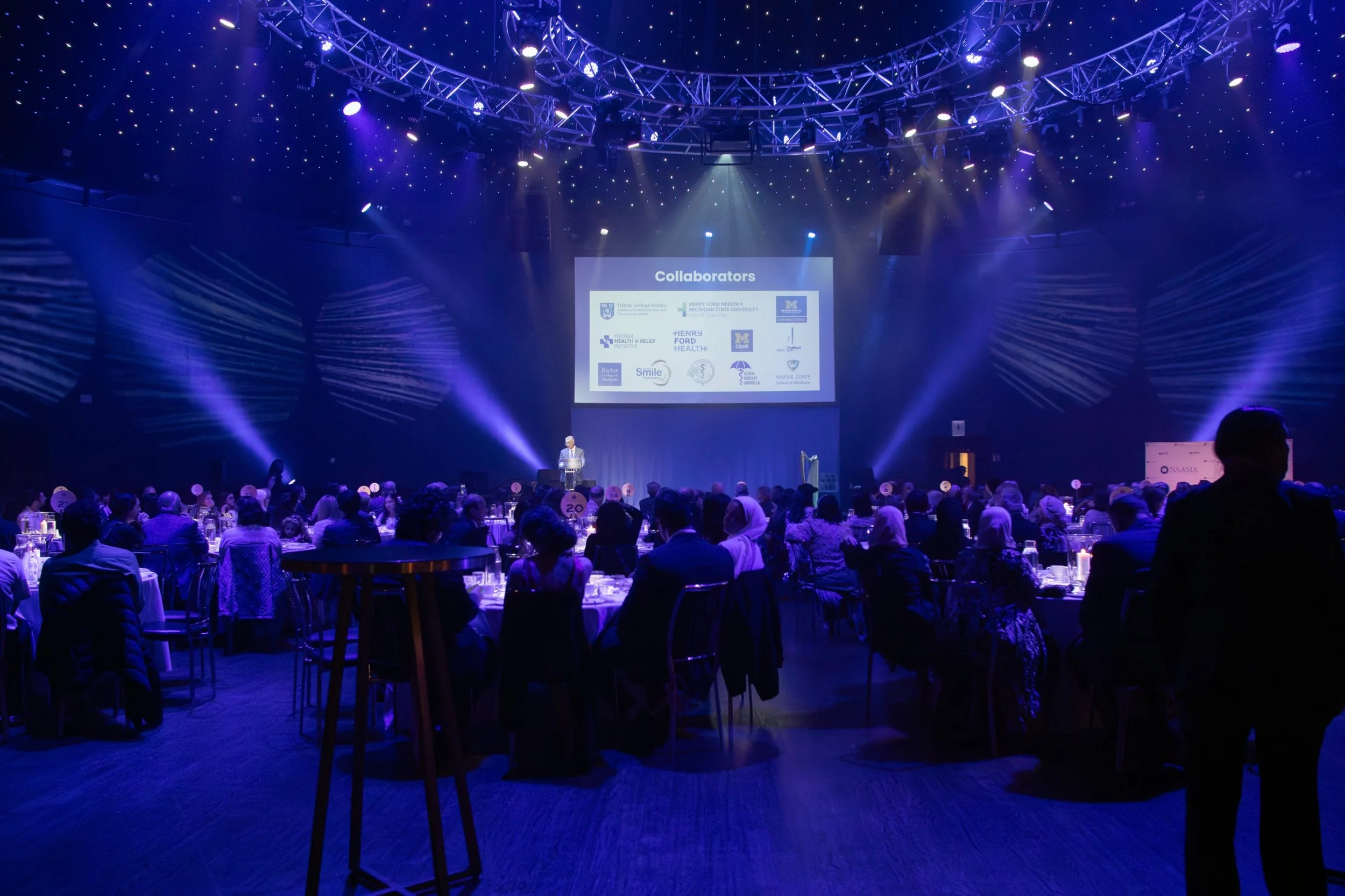 A large conference or event hall with a stage displaying a slide titled 'Collaborators' and logos of various organizations. The audience is seated at round tables, and the room is illuminated with purple and blue lighting, creating a formal atmosphere.