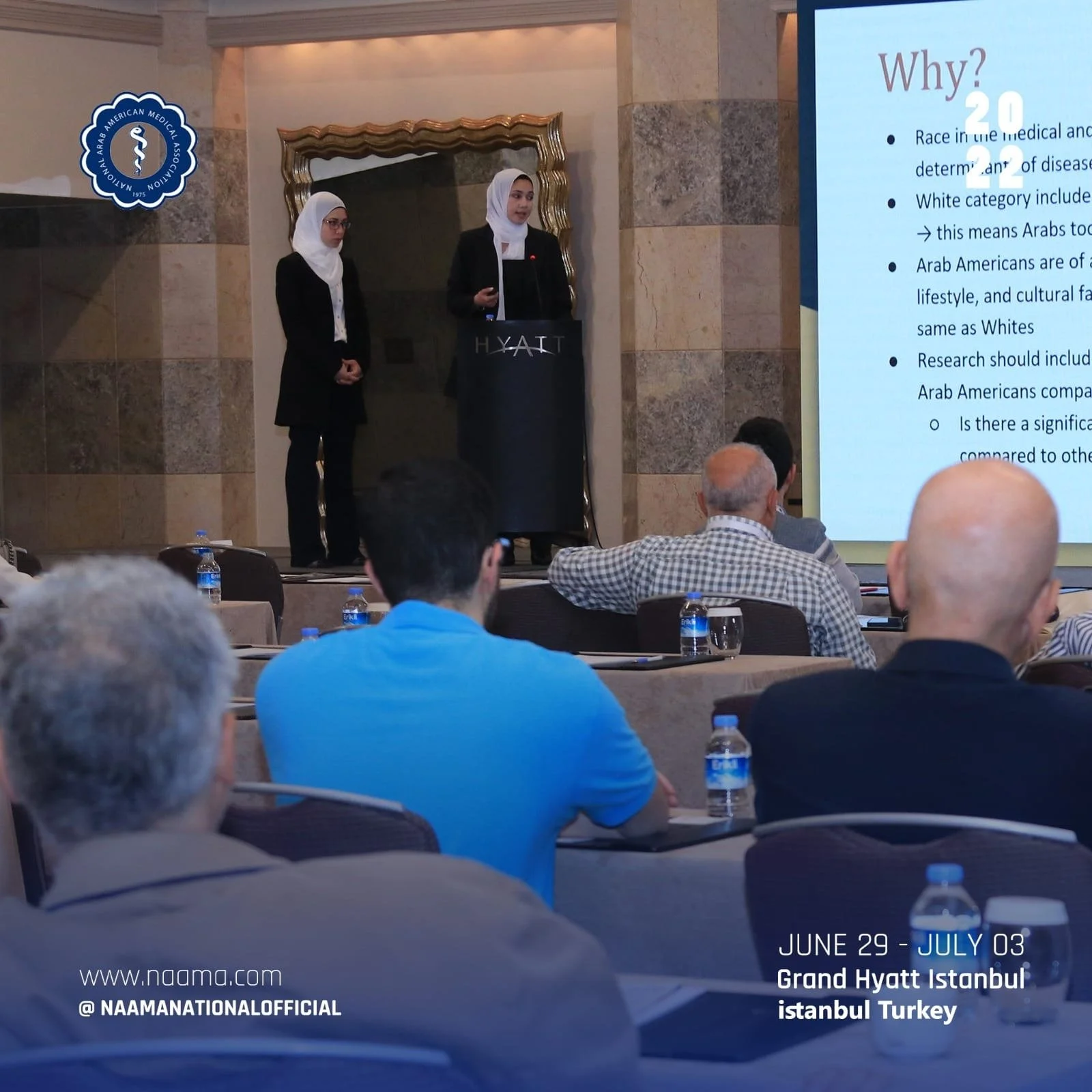A woman in a black suit and white hijab is speaking at a podium at the Grand Hyatt Istanbul hotel, with a large screen displaying a presentation slide behind her. An audience is seated, listening, with water bottles on the tables.