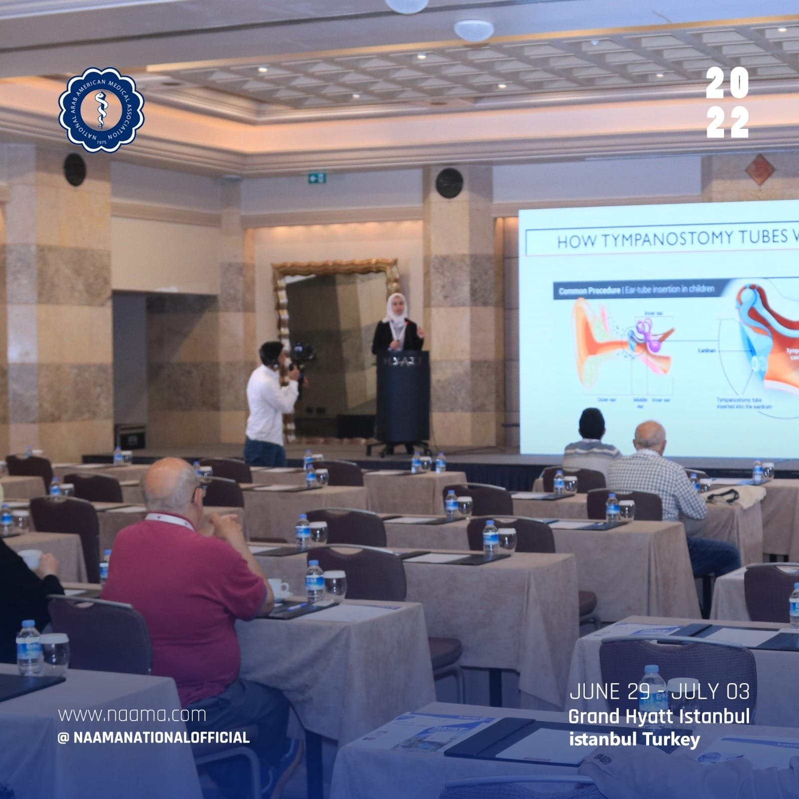 A conference room with several attendees seated at tables, some taking notes, listening to a woman giving a presentation. The presentation slide displays information about tympanostomy tubes, with an illustration of ear anatomy and insertion procedur