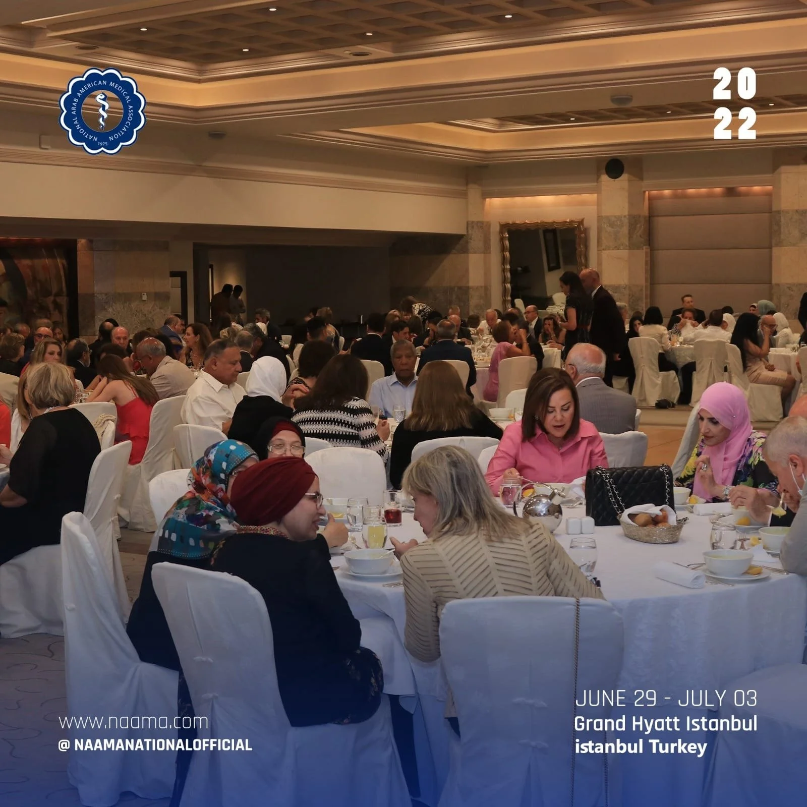 A large banquet hall filled with people sitting at round tables covered with white tablecloths. The attendees are engaged in conversation, dining, and socializing. The room has a high ceiling, beige walls, and decorative lighting. There are two promi