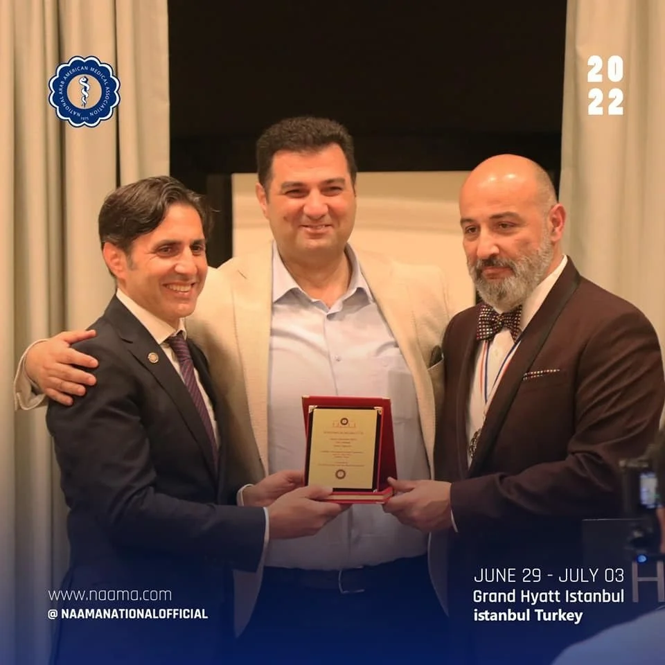 Three men are standing together, with the man on the left offering a plaque to the man on the right. The man in the center is smiling. The event is related to NAAMA, held at the Grand Hyatt Istanbul, Turkey, from June 29 to July 3, 2022.