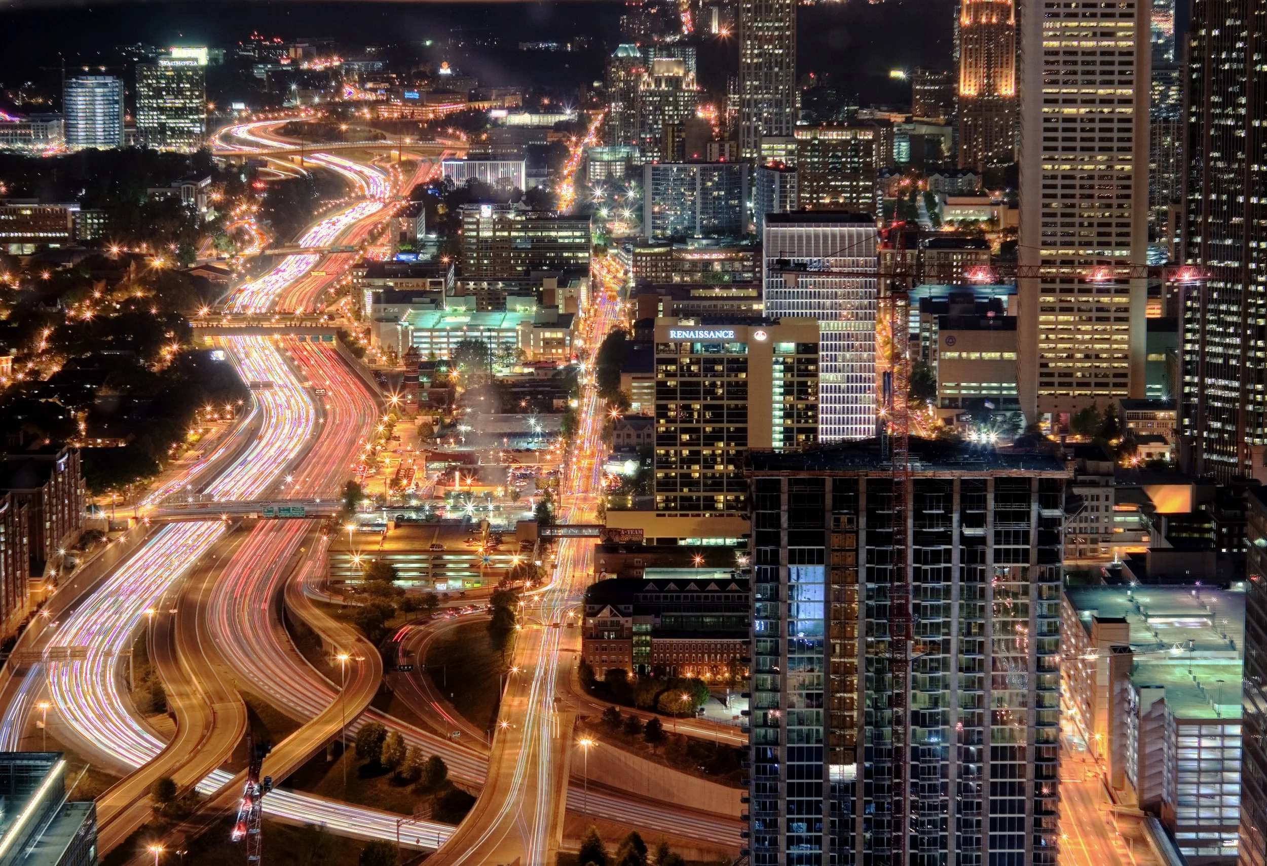 Nighttime cityscape with illuminated skyscrapers and busy highways with light trails from moving vehicles.