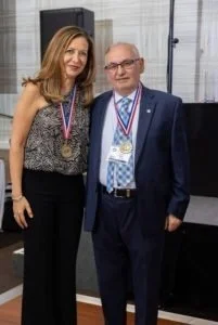 A woman and an older man standing together, both wearing medals around their necks, at an indoor event or ceremony.