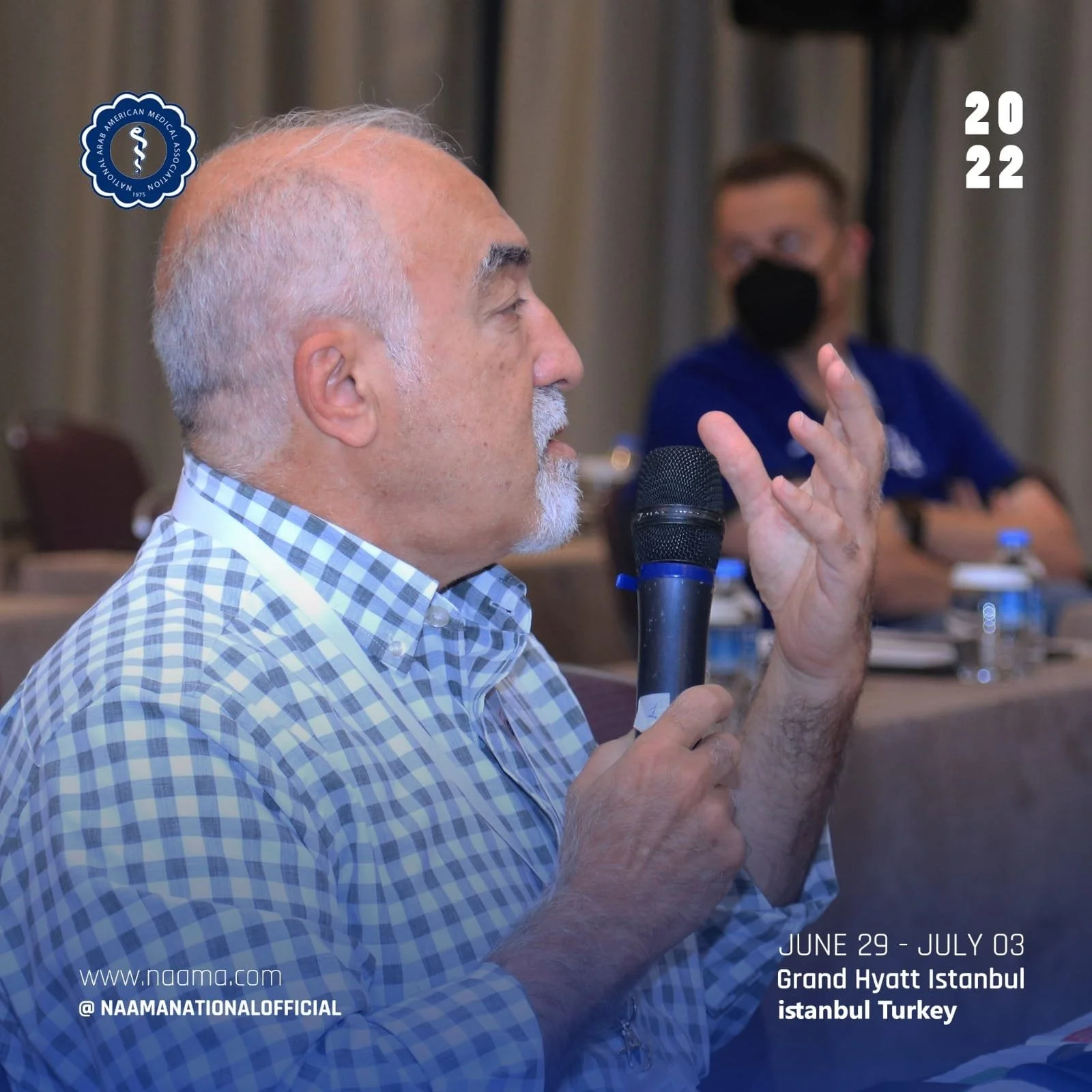 An older man with white hair and a beard, wearing a blue and white checked shirt, speaking into a microphone at a conference. In the background, a woman wearing glasses and a black face mask is sitting at a table. The event is held at the Grand Hyatt