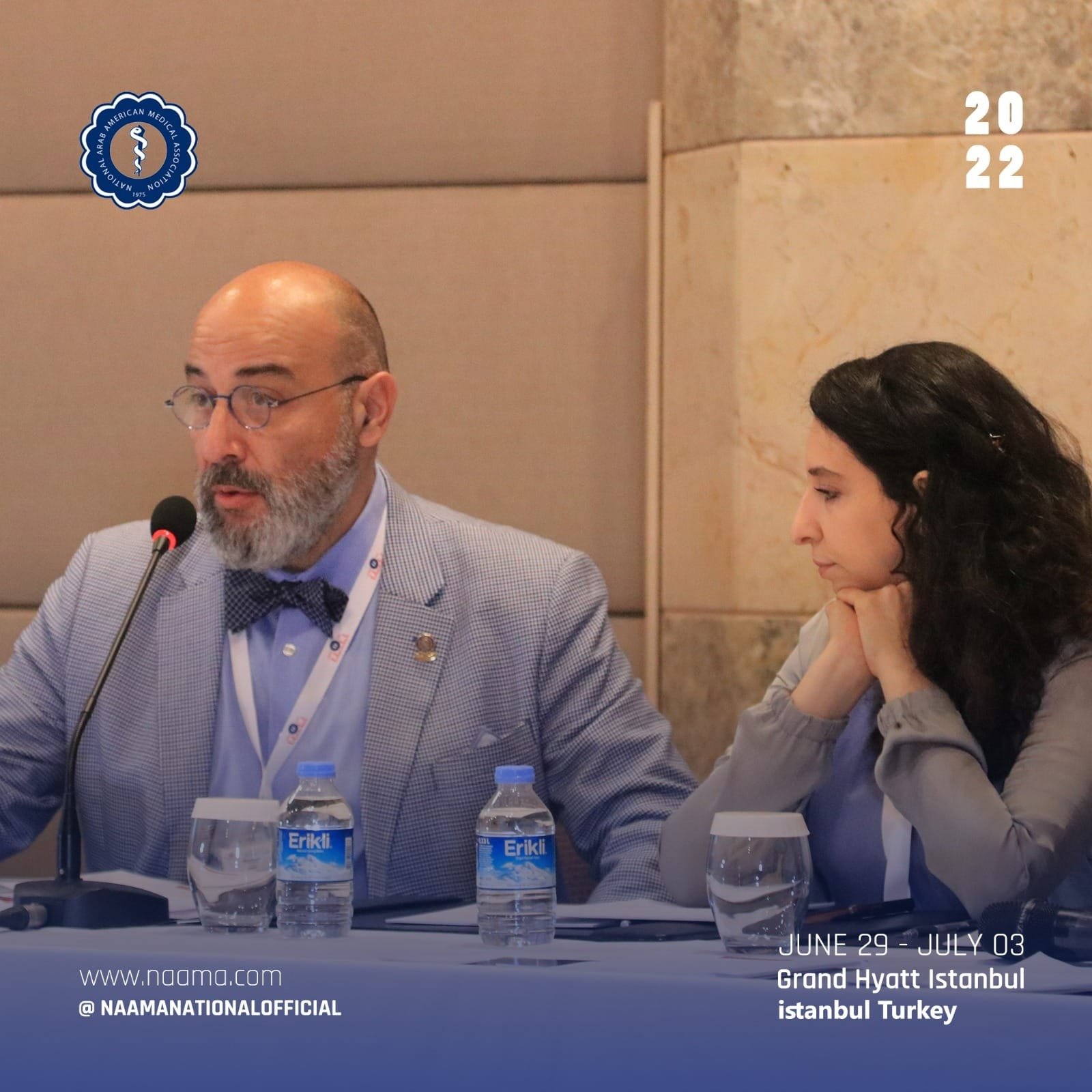 A man with glasses and a gray beard wearing a light gray suit and bowtie, and a woman with dark curly hair wearing a light gray blazer, sitting at a table with microphones and water bottles, during a conference at the Grand Hyatt Istanbul, Turkey, fr