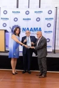 Three people at a podium during a NAMA event, with two women and one man, exchanging a handshake and holding a trophy or award, in front of a NAMA branded backdrop.
