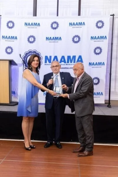 Three people standing on a stage, two men and one woman, during an award presentation or ceremony at NAMA, with a backdrop displaying the NAMA logo and website.