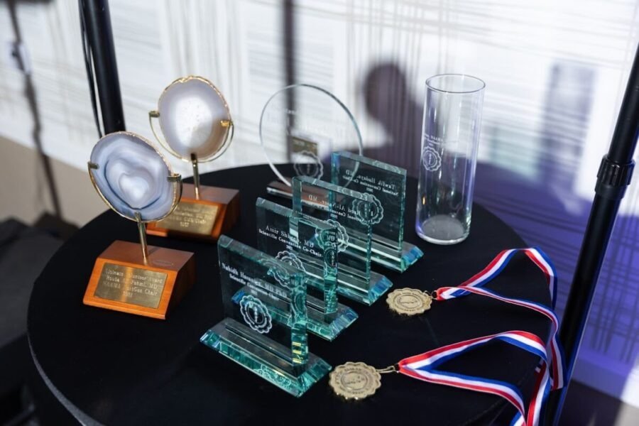 Awards and medals displayed on a black table, including engraved plaques, crystal trophies, and medals with ribbons, with a blurred background.