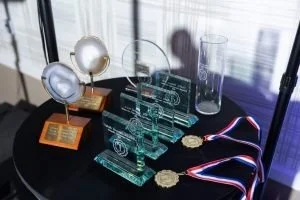 Trophies and medals on a black table with a transparent slide showing technical drawings and a glass of water in the background.