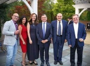 Group of six professionally dressed people standing outdoors in front of green trees and a white structure, posing for a photo.