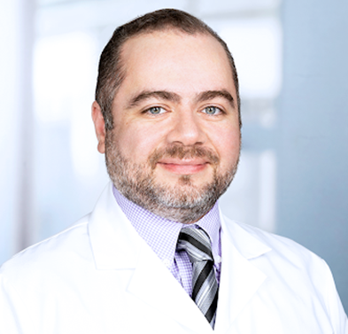 A male doctor with a beard and short hair, wearing a white coat and a striped tie, standing in a bright clinical environment