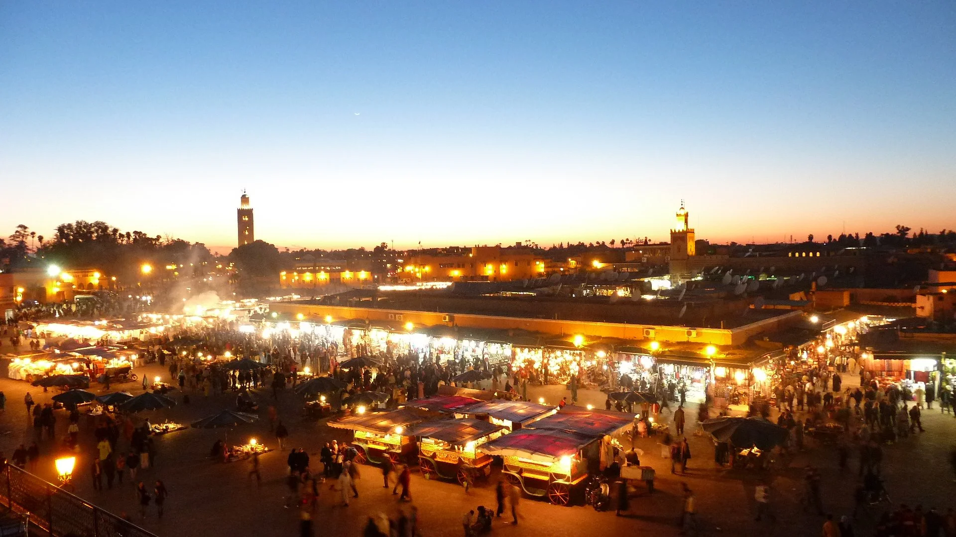 A lively market scene at sunset with numerous stalls, lights, and a crowd, and a skyline featuring tall towers and buildings in the background.