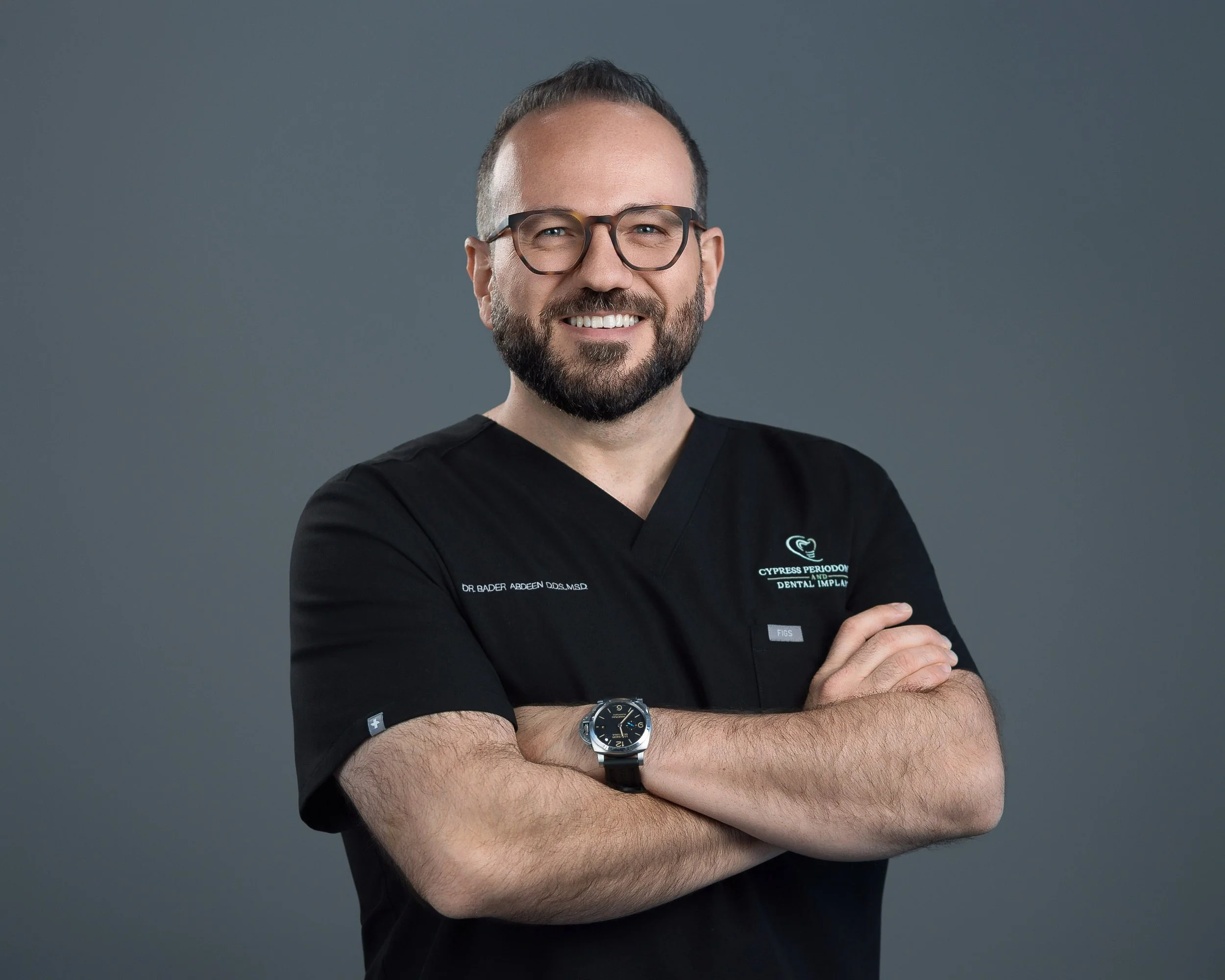 A man with short dark hair, a beard, and glasses, wearing a black medical uniform with a name tag that reads "Dr. Bader Abdeen DDS, MSD" and a wristwatch, standing against a plain gray background.