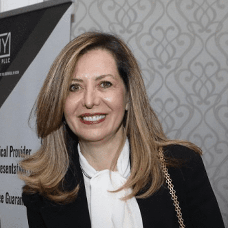 A smiling woman with shoulder-length light brown hair, wearing a black blazer and white blouse, standing indoors near a decorative wallpaper background.