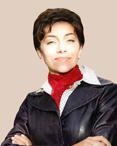 Woman with short dark hair wearing a black leather jacket, white polka dot shirt, and red scarf, standing against a beige background.
