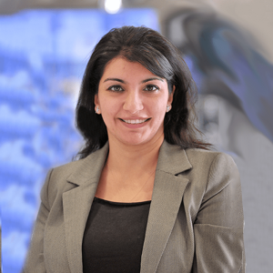 A woman with dark hair wearing a gray blazer and black top, smiling in front of a blurred background.