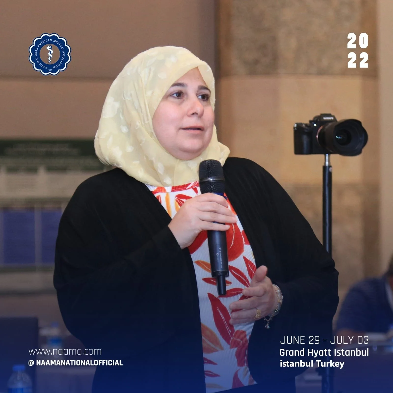 A woman wearing a light yellow headscarf, red patterned dress, and black cardigan holding a microphone. A camera on a tripod is in the background. The image is from NAAM, taking place from June 29 to July 3 at the Grand Hyatt Istanbul in Turkey, with