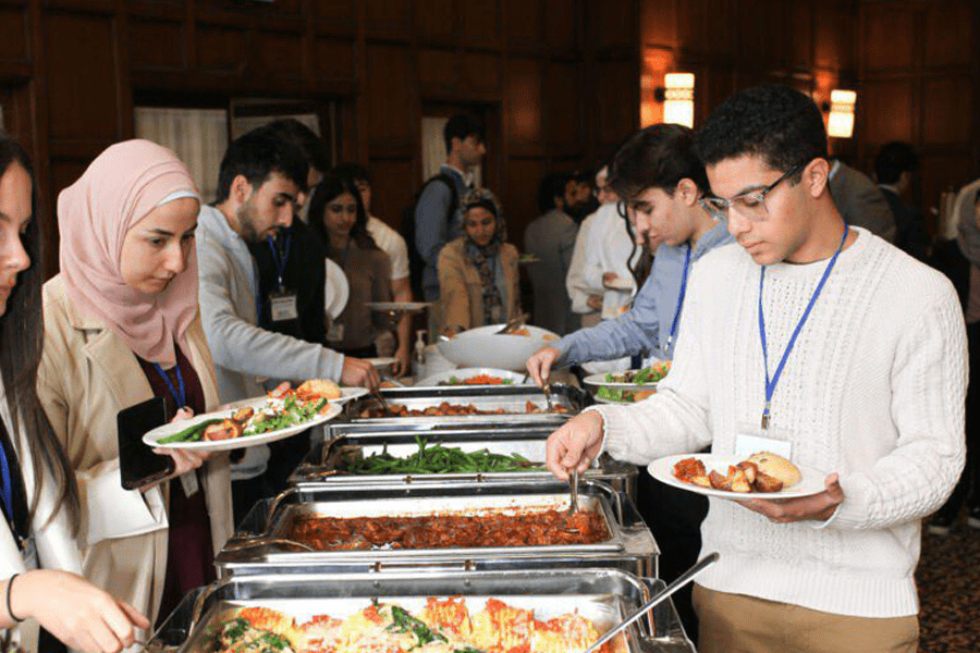 People at a buffet line selecting food, with various dishes in chafing dishes and individuals holding plates in a dining hall setting.