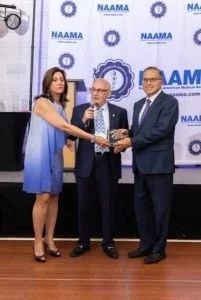 Three professionally dressed individuals standing on a stage, with a NASMA and other organizational banners in the background. The two men in the center are holding an award or plaque, and the woman on the left is reaching towards it.