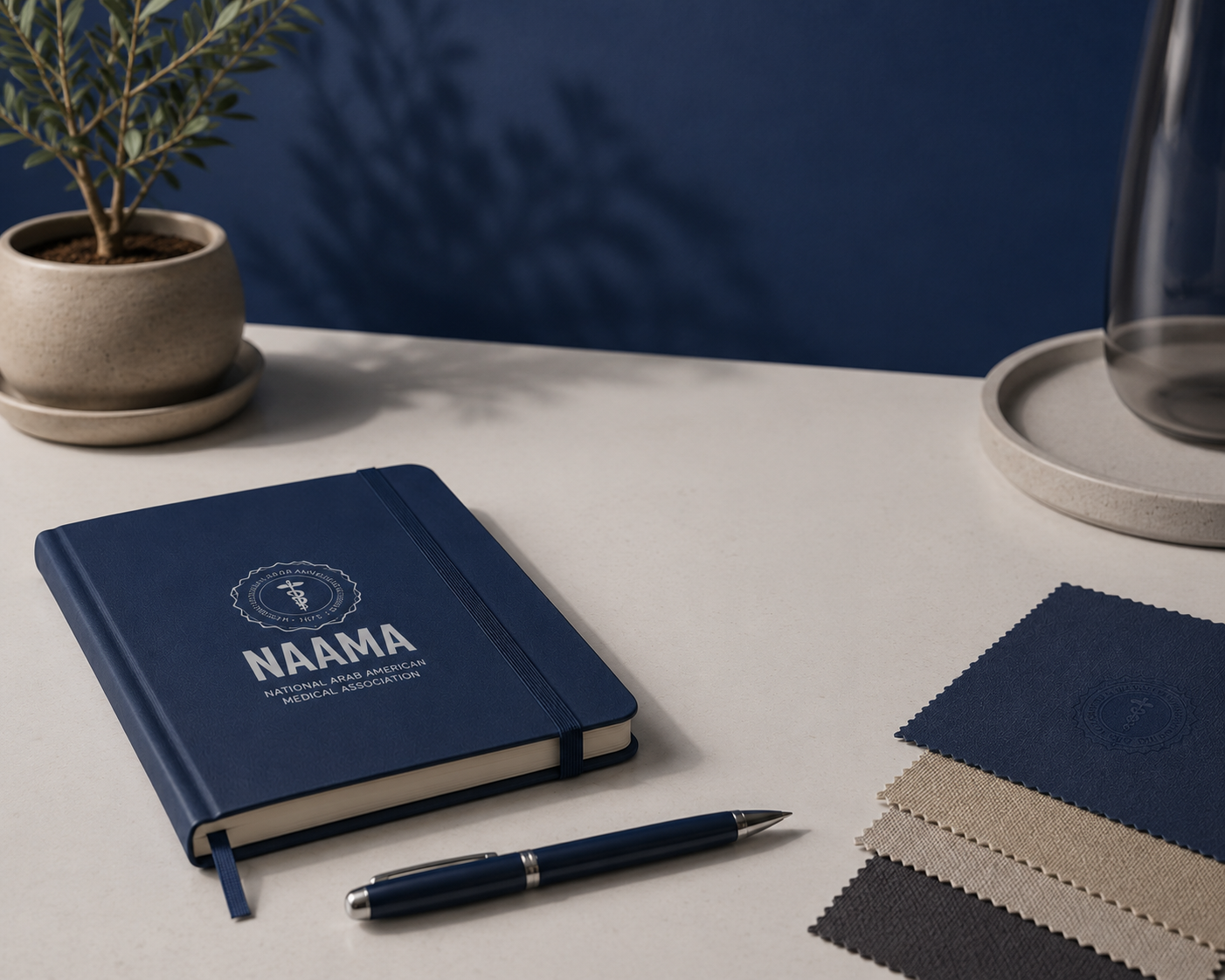 A desk with a blue notebook labeled 'NAAMA' for the National Arab American Medical Association, a black pen, fabric swatches in beige, blue, gray, and black, a potted plant, and a glass vase on a tray. The background is a blue wall.