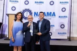 Three people posing for a photo at an award ceremony, one holding a glass and trophy, with a NAAMA backdrop.