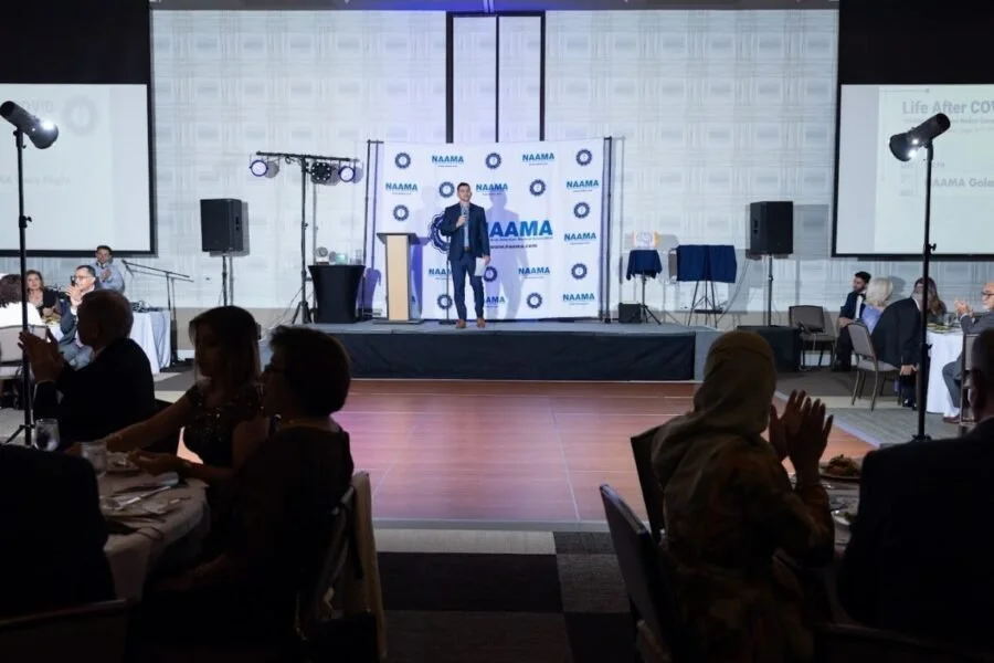 Woman speaking on stage at a NAAMA event while attendees sit at round tables in a banquet hall.