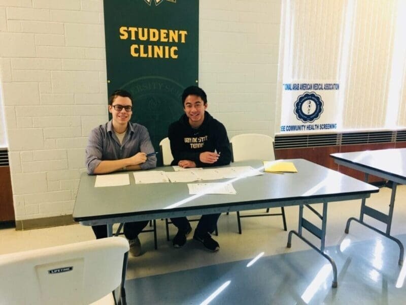Two young men sitting at a table during a student clinic event, smiling and surrounded by papers, in a room with banners that say 'Student Clinic' and health association.
