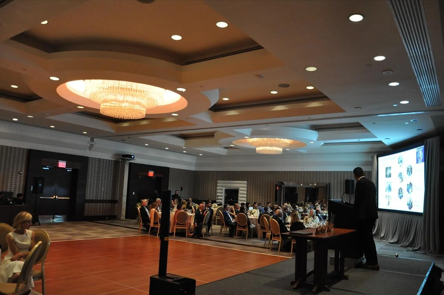 A banquet hall or conference room with guests seated at round tables and a speaker at a podium. There is a large screen displaying presentation slides on the right and chandeliers hanging from the ceiling.